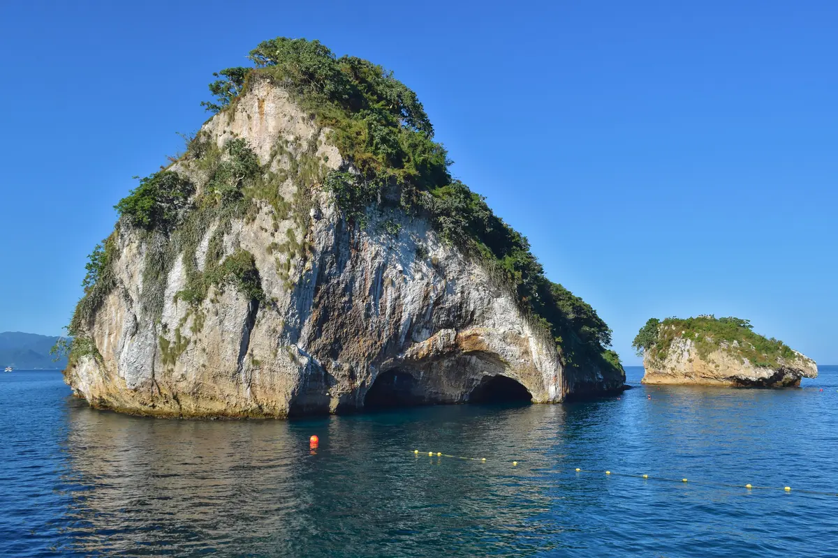 Two rounded rock islands rising from calm blue water under a clear sky, one with a sea arch at its base (the Los Arcos rocks off Mismaloya, on the bay's southern coast)