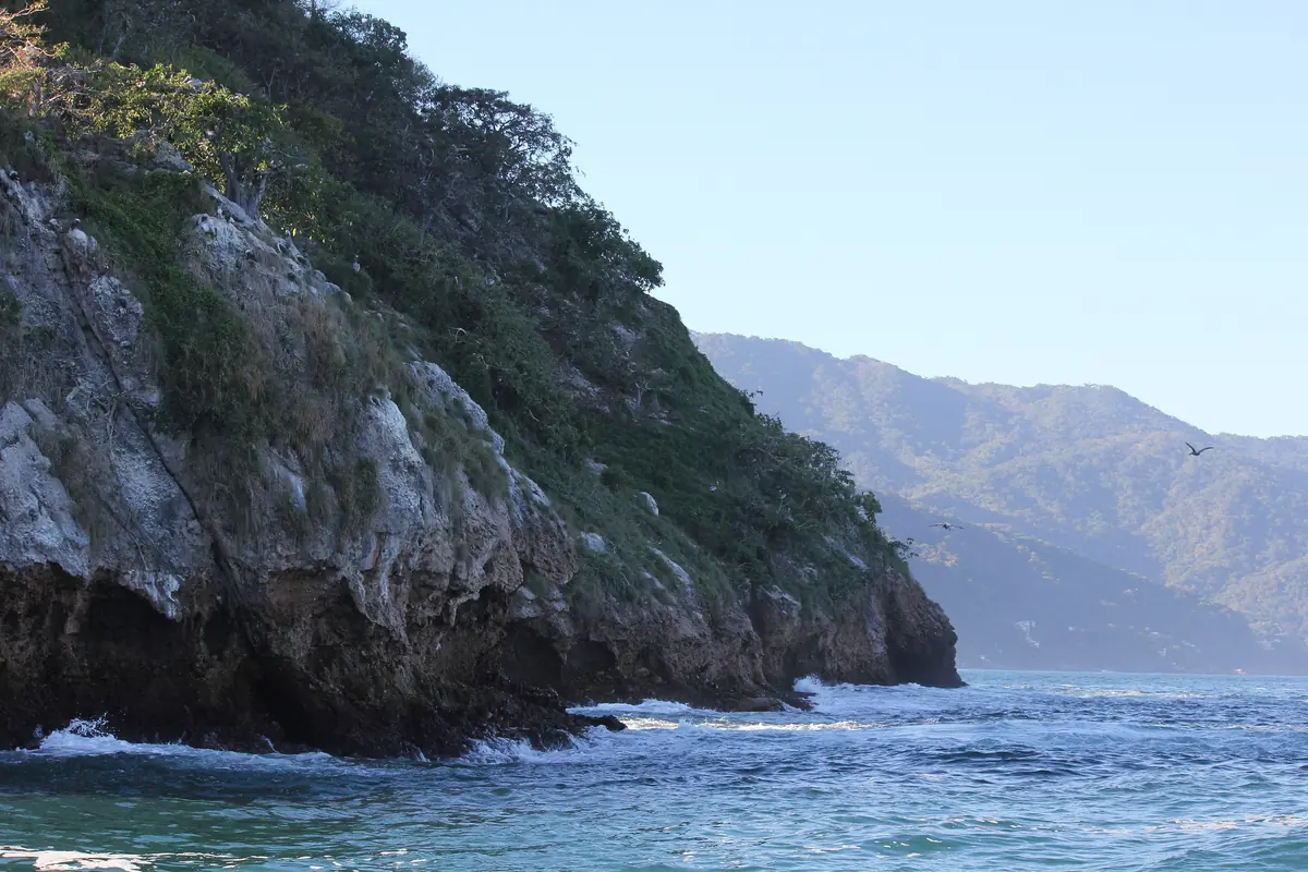 A steep green headland dropping to blue water, with a hazy mountain coast beyond (a headland on the boat route toward Las Caletas, not the cove itself)