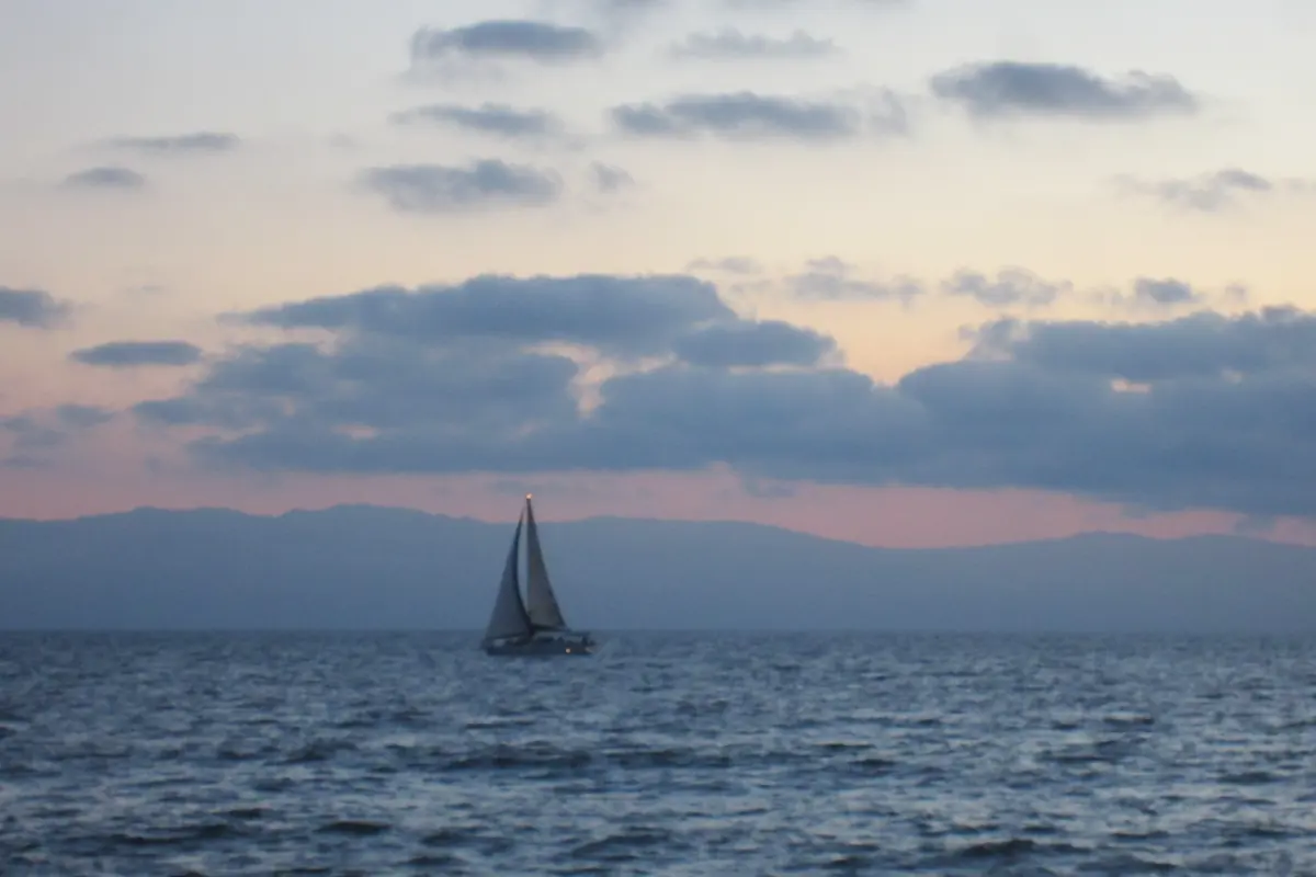 A single sailboat on dark blue water under scattered grey clouds, with a hazy mountain coast and a faint pink glow at dusk (a sailboat on Banderas Bay, not a show boat)